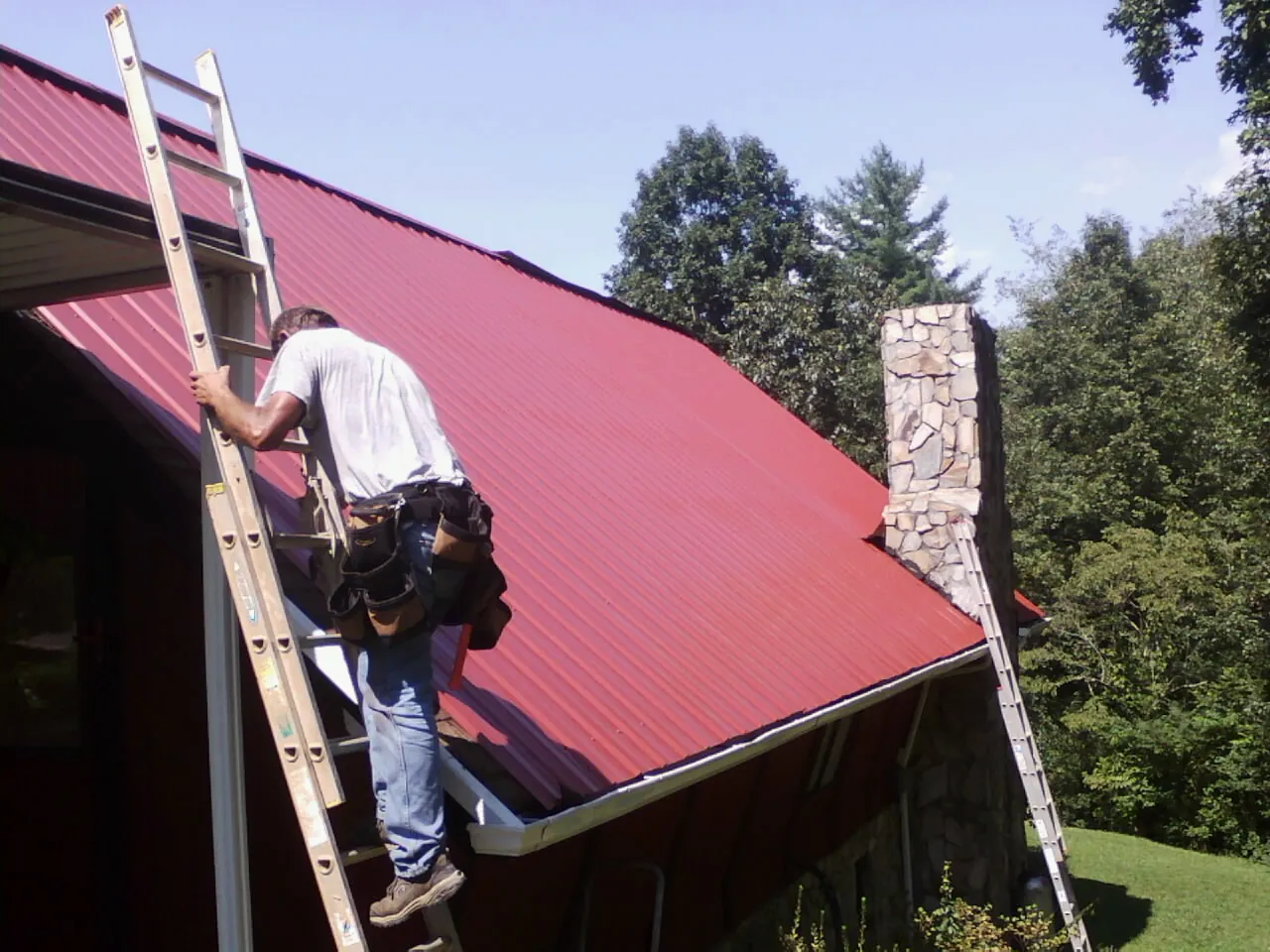 Roofer on a ladder beside a red metal roof and stone chimney — Soffit & Fascia Repair by Roofing Brothers in Fe Warren Afb